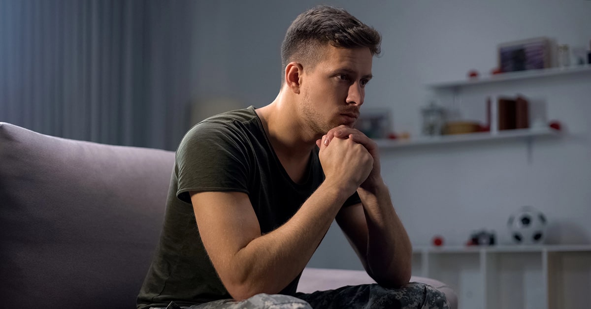 Man sitting on couch in dim room, hands clasped, staring ahead.