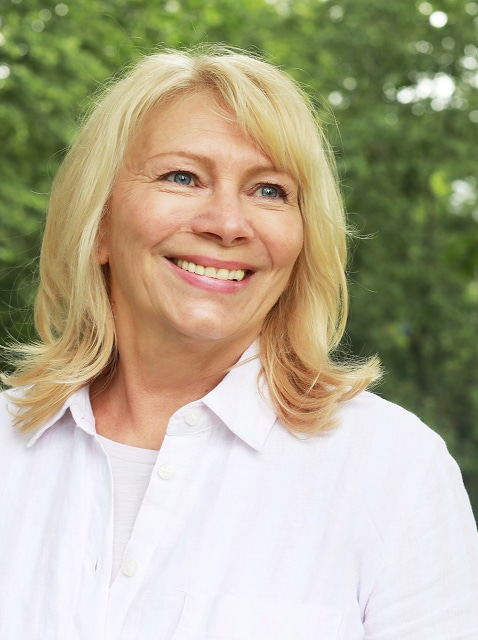 Smiling blonde woman in a white shirt outdoors with blurred green trees