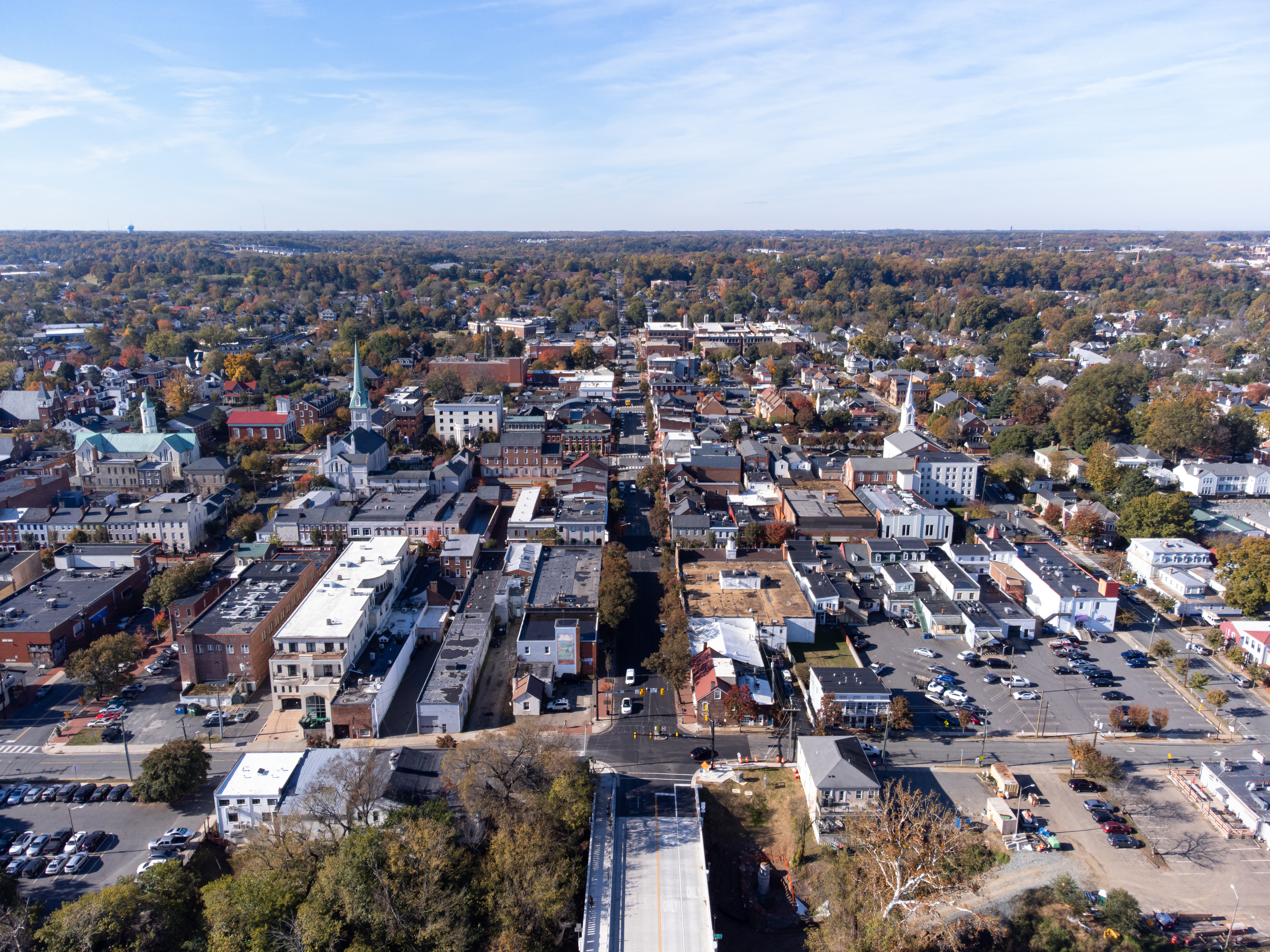 Aerial view of historic downtown Fredericksburg on a clear autumn morning, featuring tree-lined streets, brick buildings, church steeples, and a small-town urban layout.